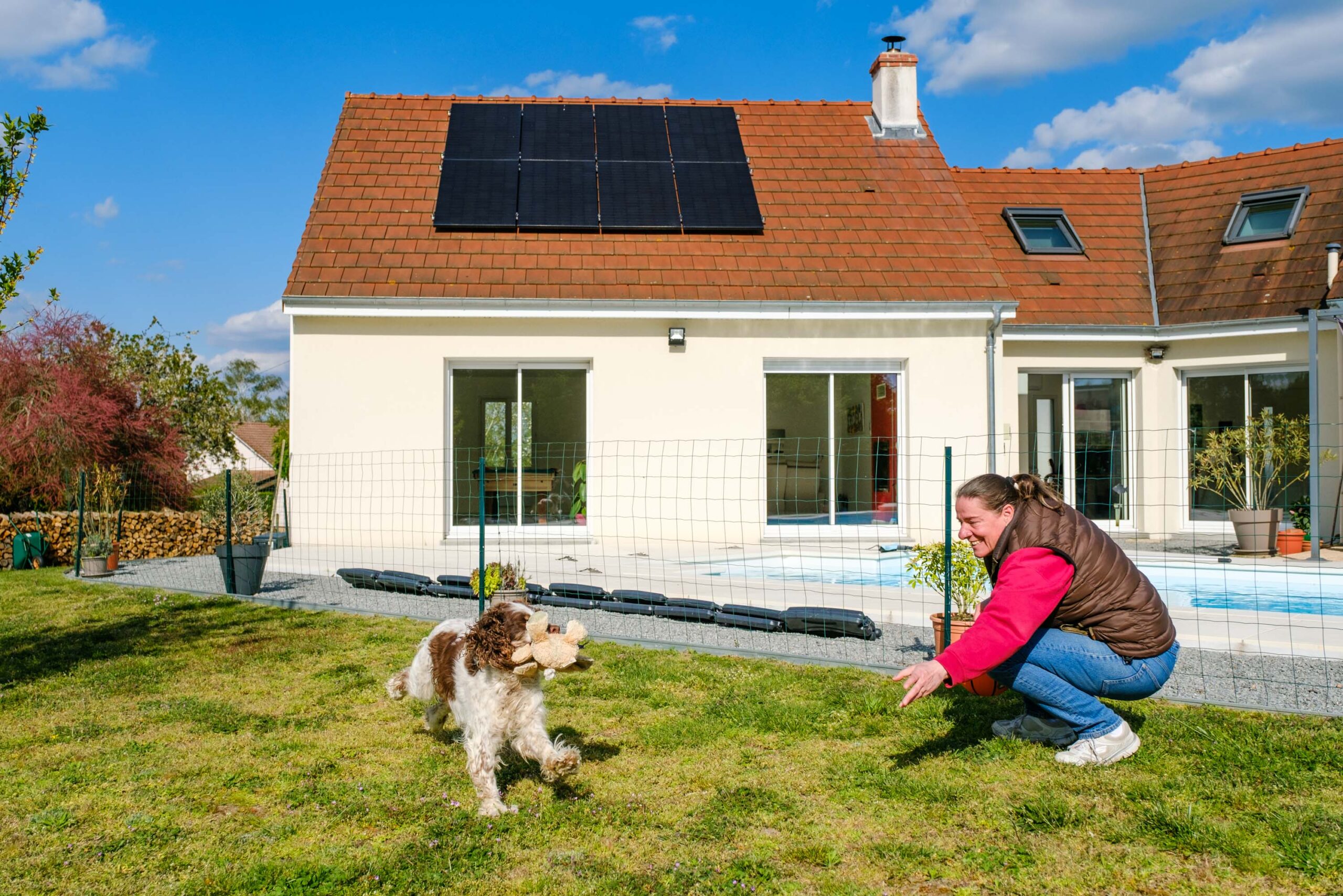 femme jouant avec son chien dans son jardin, avec des panneaux solaires sur le toit de sa maison en fond